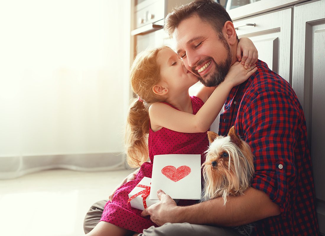 Personal Insurance - Daughter Hugs and Kisses Her Father While He Holds a Small Dog at Home